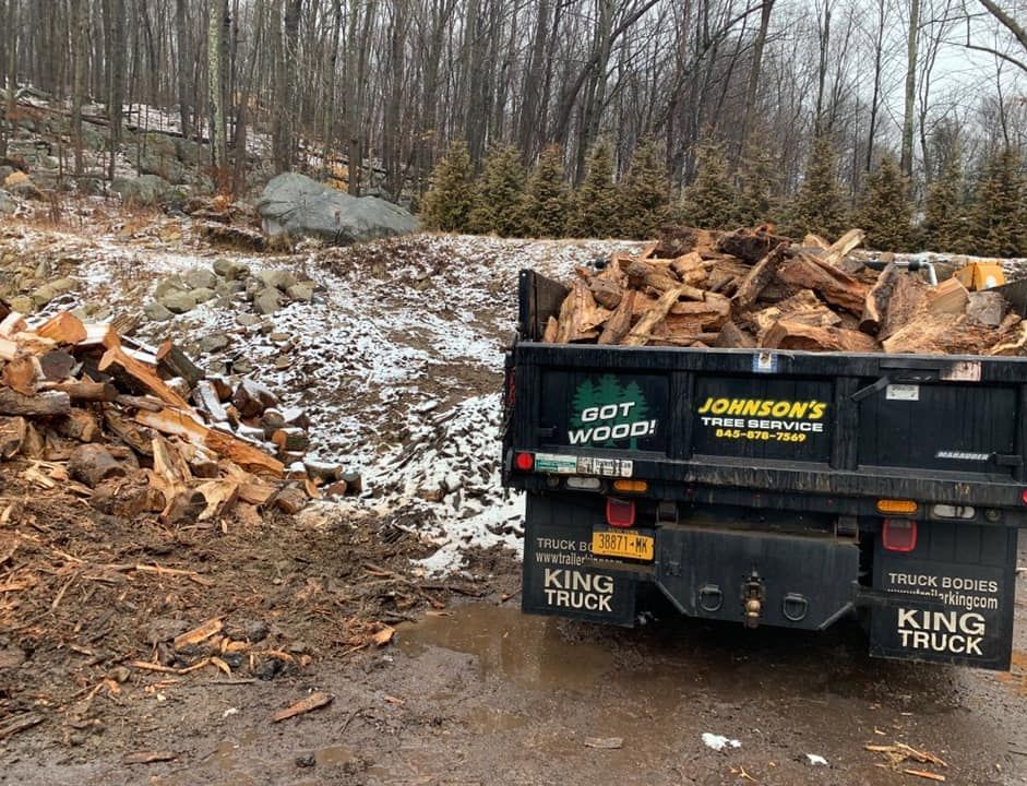 Black dump trailer loaded with firewood in a snowy wooded clearing, with split logs stacked nearby.