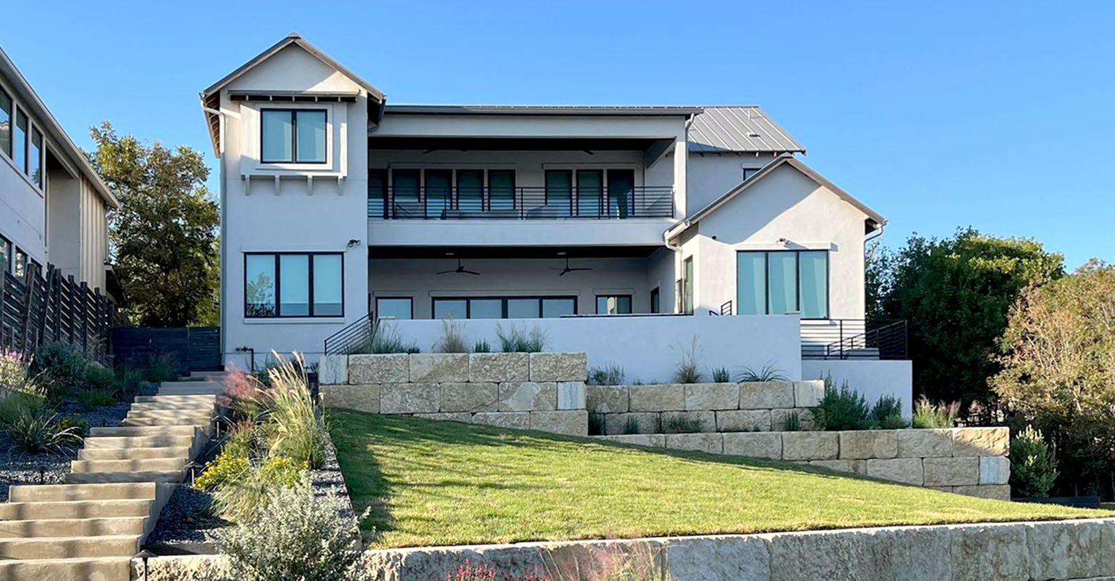 Two-story house with light gray stucco, balcony, and stone landscaping on a bright day.