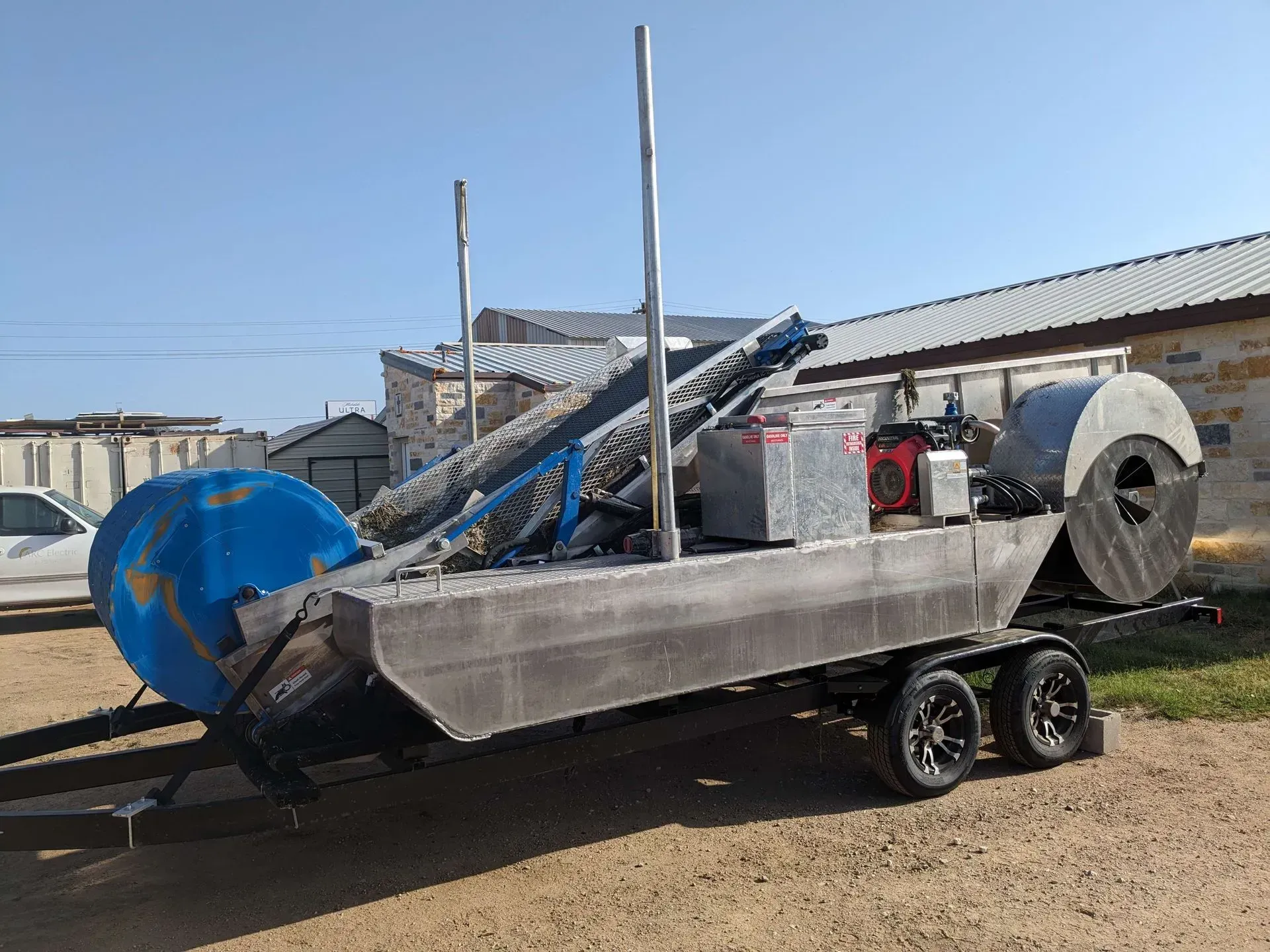 Gray metal watercraft on a trailer with a blue drum, conveyor belt, and engine. Outdoors.
