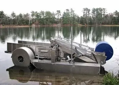 Floating metal debris skimmer in a lake, removing surface waste; trees reflected in the water.