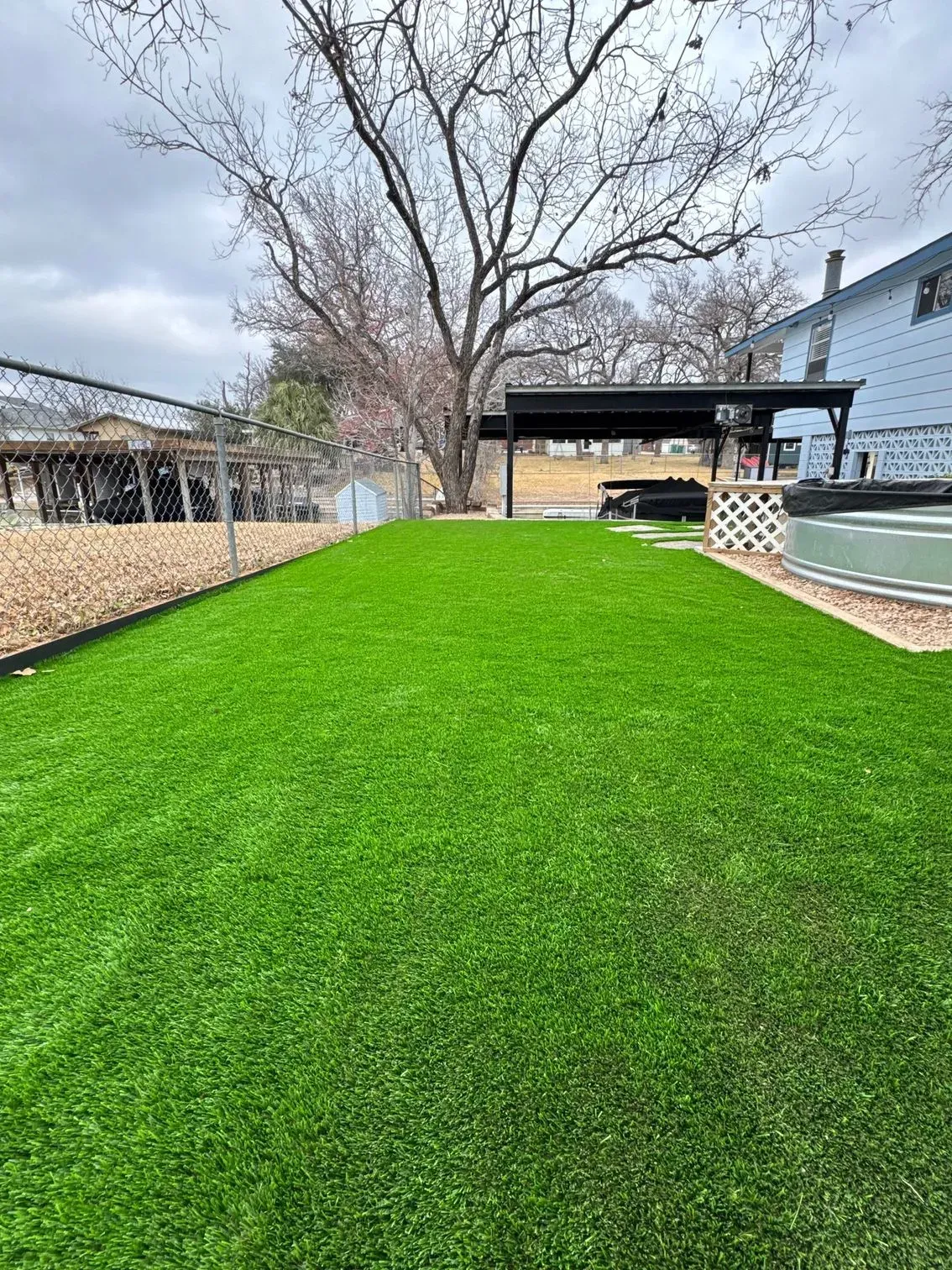 Bright green artificial turf in a backyard with a covered patio, fence, and bare tree.