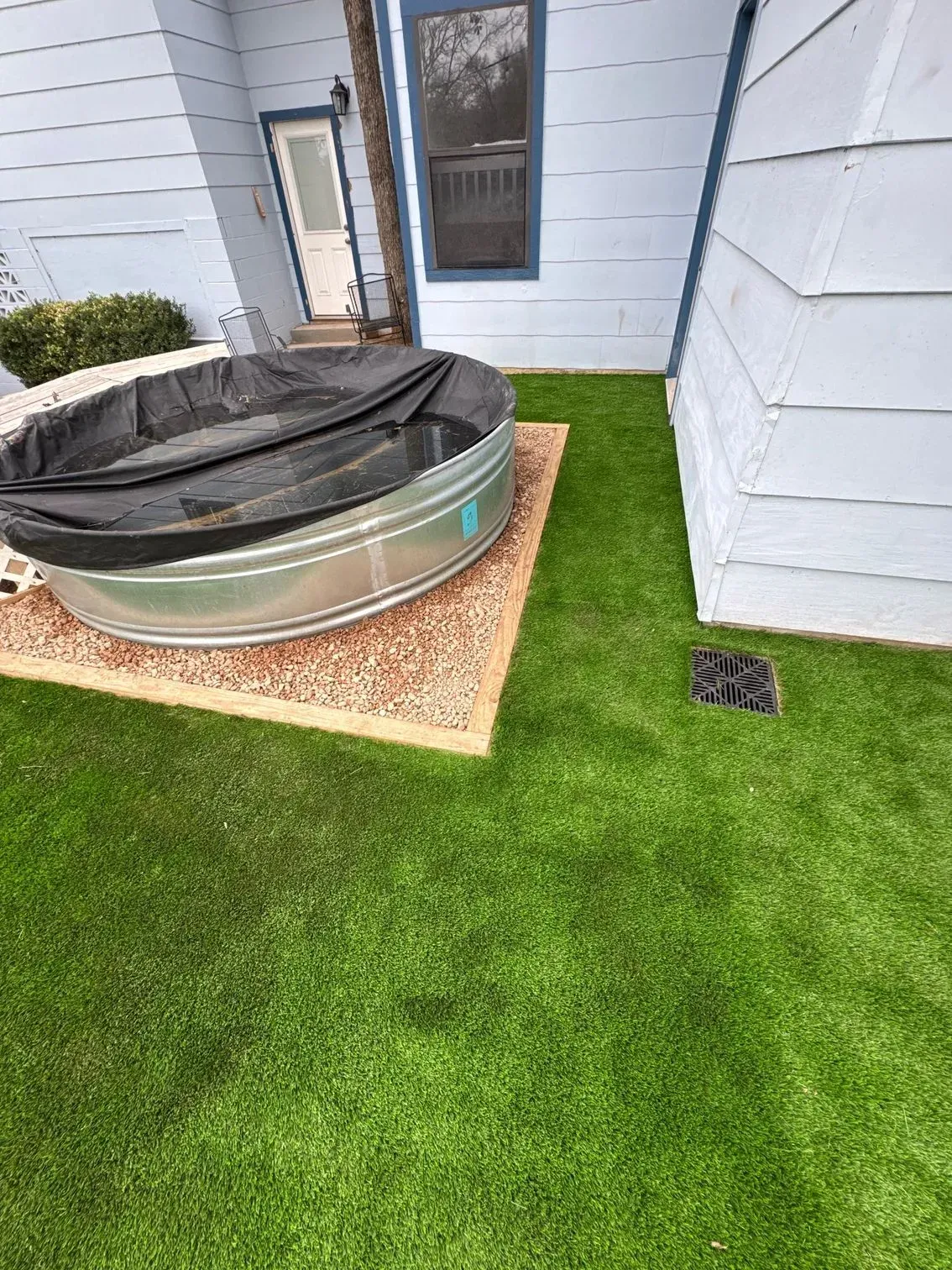 Backyard with a metal stock tank covered by a black tarp, surrounded by artificial grass.