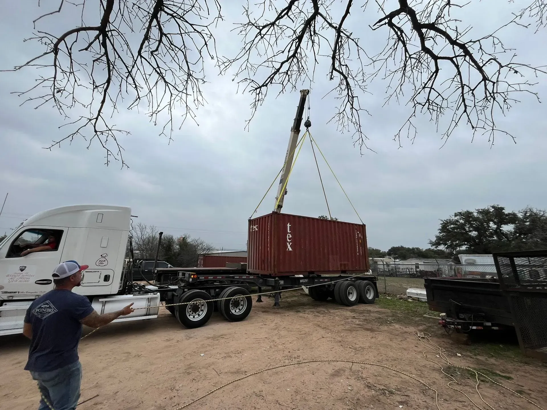 A man supervises a crane lifting a red shipping container off a trailer truck on a cloudy day.