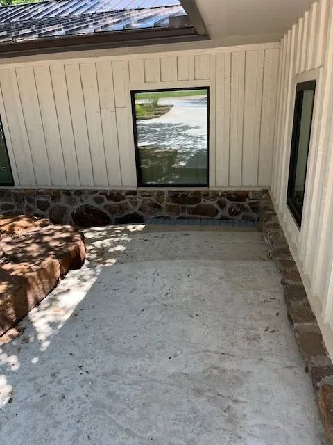 Exterior view of a house with concrete patio, stone foundation, white siding, and black-framed windows.