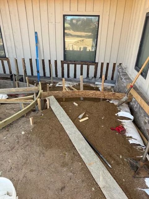 Construction site with wooden forms and gravel base, near a house with a window.