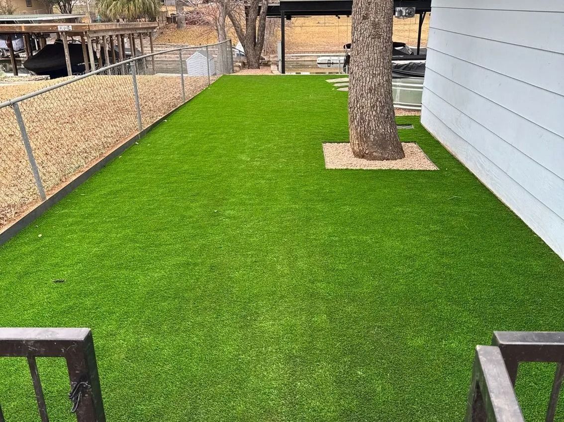 Green artificial turf in a backyard with a tree and a white wall, bordered by a fence.