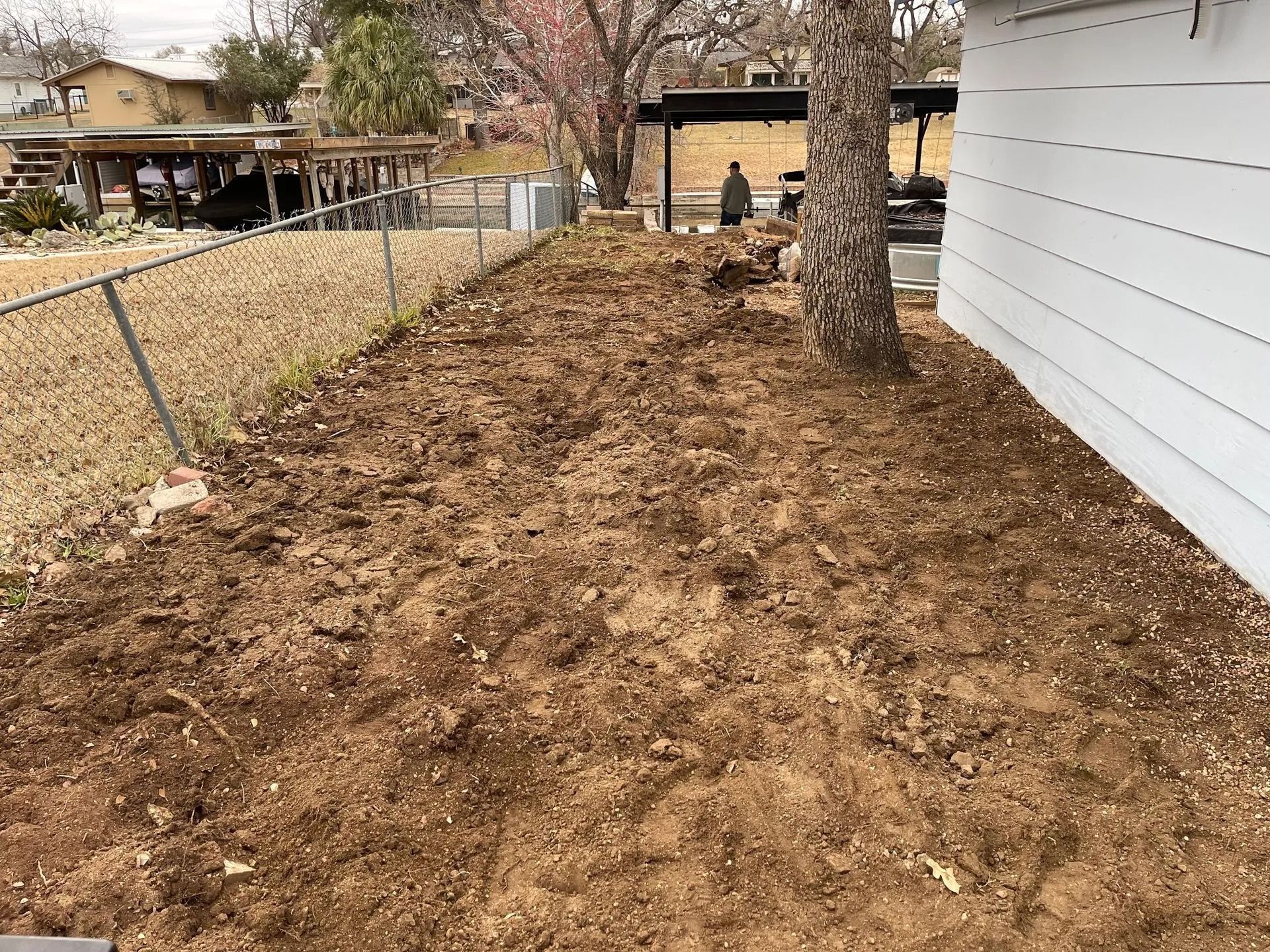 Dirt bed next to a white house and chain link fence, next to a tree, in a backyard.