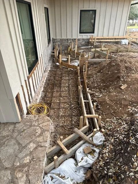 Construction site: Forms and rebar prepared for a concrete walkway next to a building with windows.