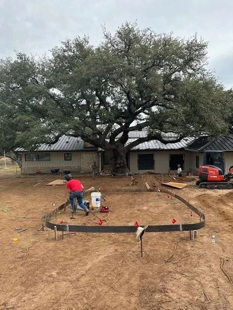 Construction site with person working on curved concrete form; house, tree, and excavator in background.