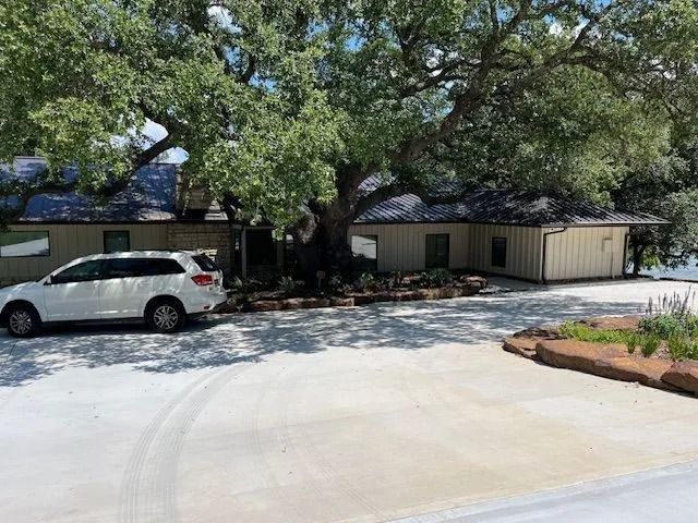 White SUV parked in front of a tan building with a large tree. Concrete driveway.