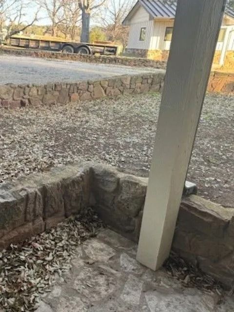 Stone patio with a white pillar, dry leaves, and a low stone wall. A trailer and small building are in the background.
