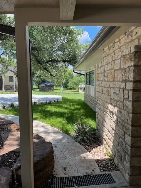 Stone-walled house with a grassy lawn, trees, and a walkway viewed from a covered porch.