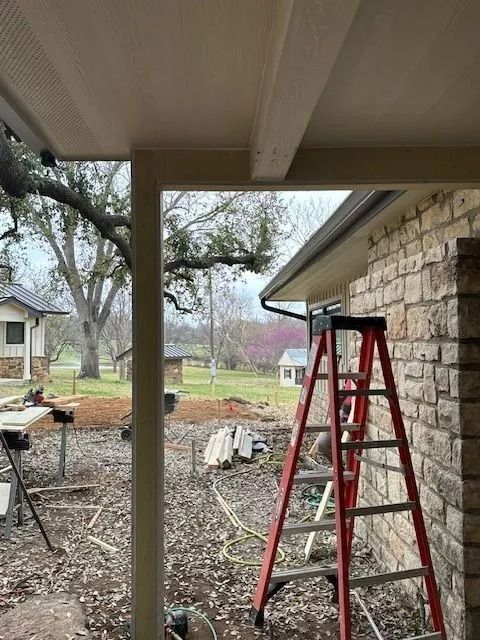 View of backyard construction from a covered porch, red ladder, brick wall, overcast sky.