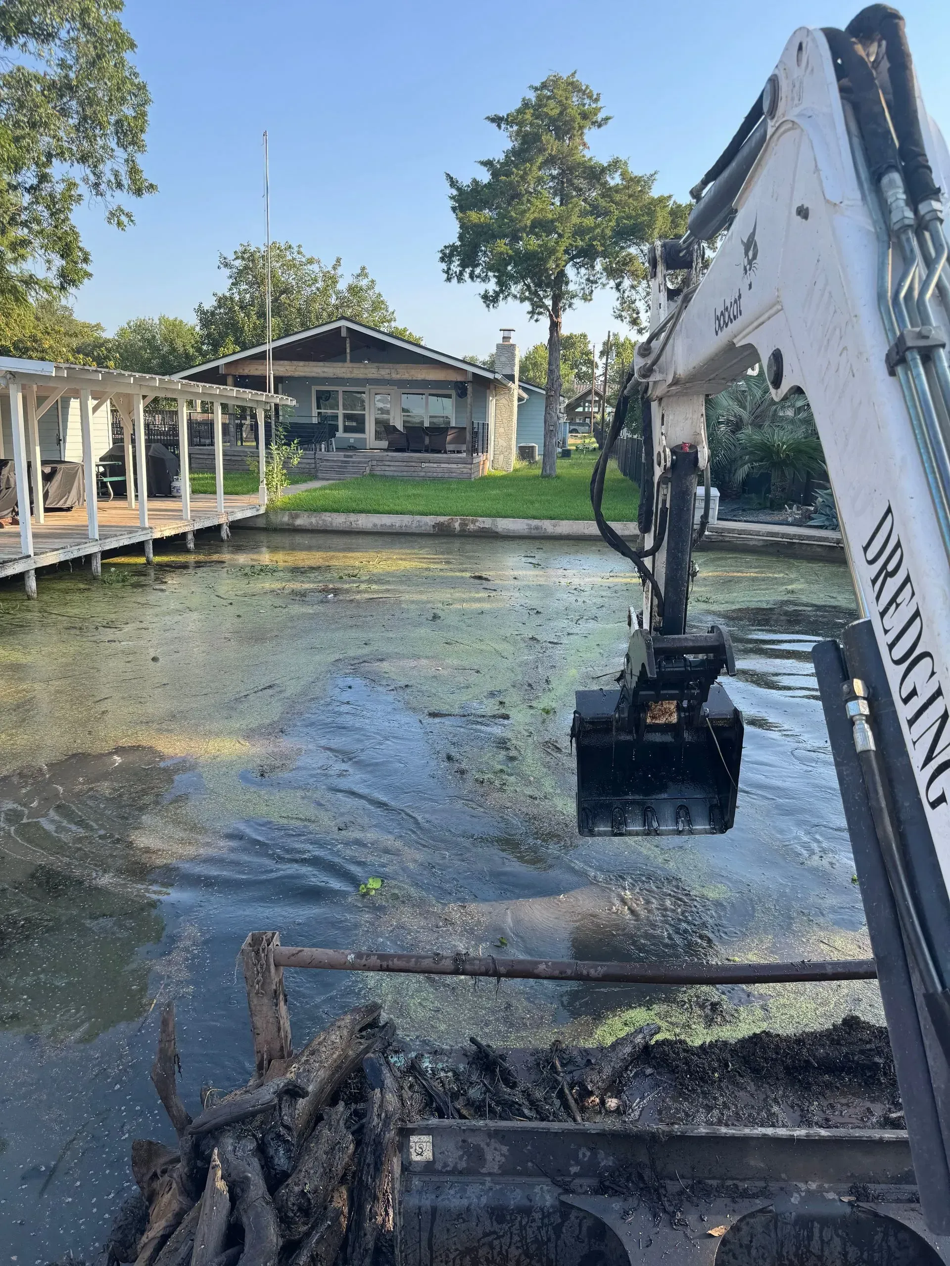 Excavator removing aquatic vegetation from a waterway near a house with a dock.