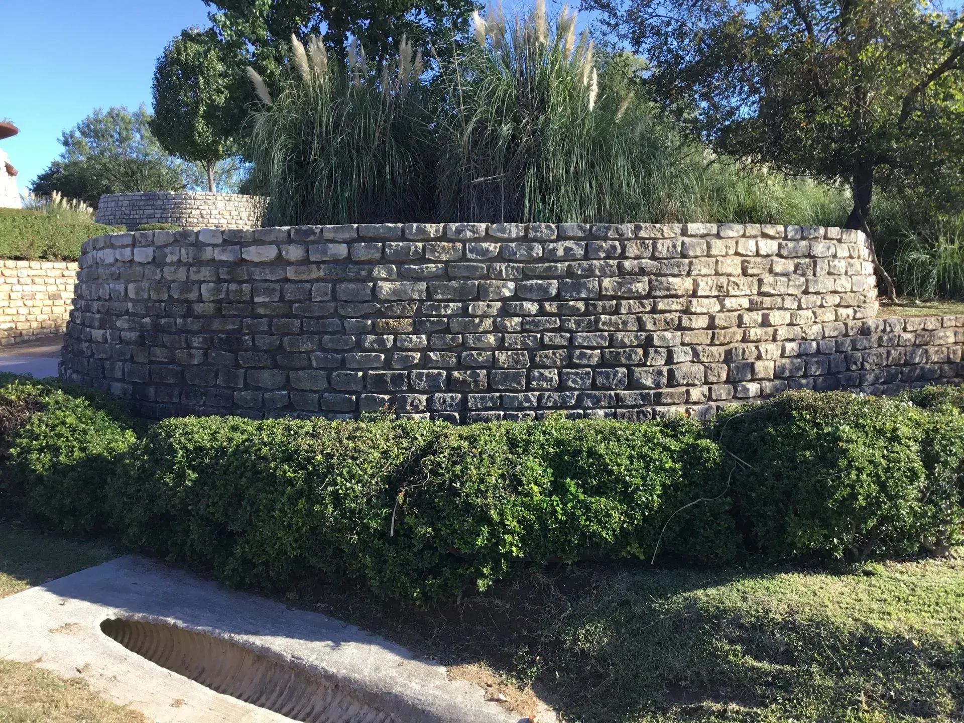 Stone retaining wall with greenery and tall grass, set in a landscape with a sidewalk.
