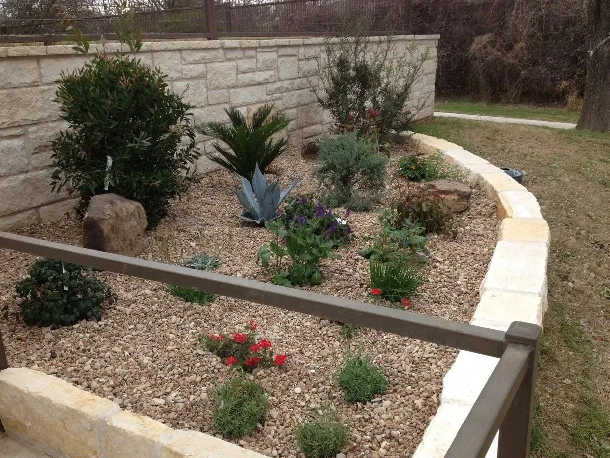 A garden bed with various plants, rocks, and wood chips, bordered by a stone wall and a brown railing.