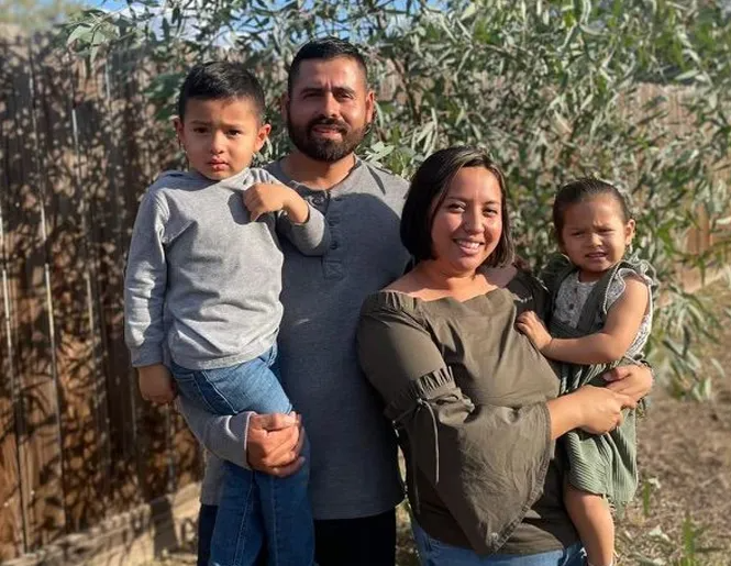 Family of four smiling outdoors: two parents holding young children.