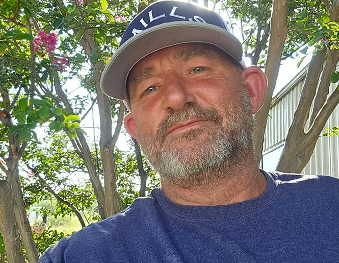 Man with a beard, wearing a baseball cap and blue shirt, looking at the camera outdoors.