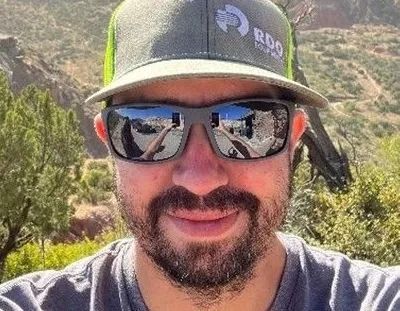 Man with sunglasses and a green hat, smiling, outdoors with a mountainous backdrop.
