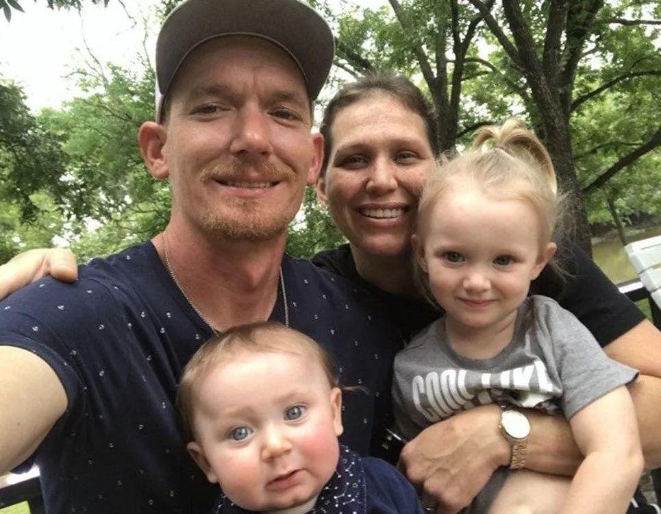 Family of four smiles for the camera outdoors; two adults and two young children.