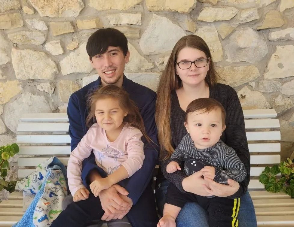 Family of four sitting on a bench against a stone wall: two adults and two young children.