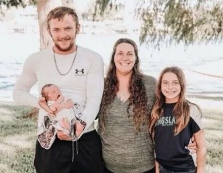 Family of four posing outdoors; father holding a newborn, mother and daughter smiling.