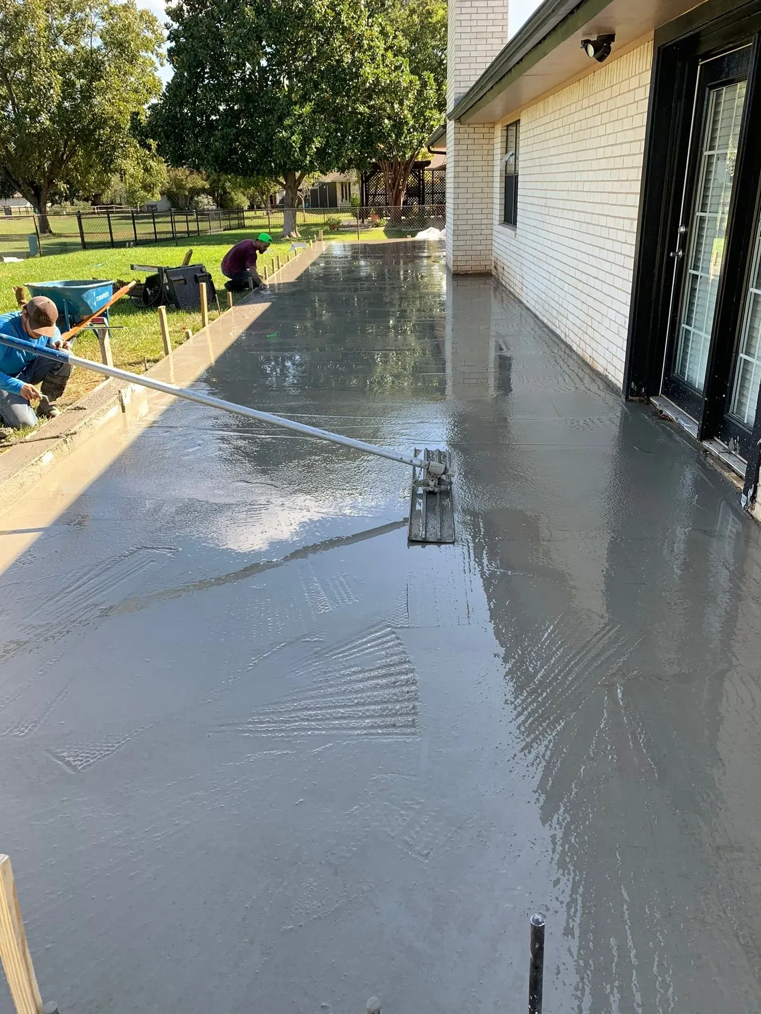 Workers smoothing freshly poured gray concrete patio next to a house, sunny outdoor setting.