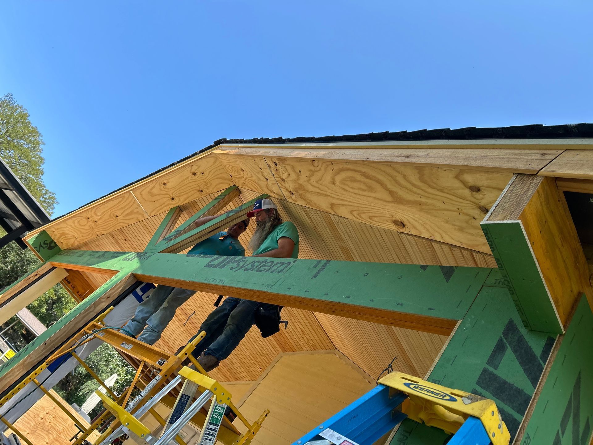 Two people are constructing a roof on a building, using a ladder and scaffolding.