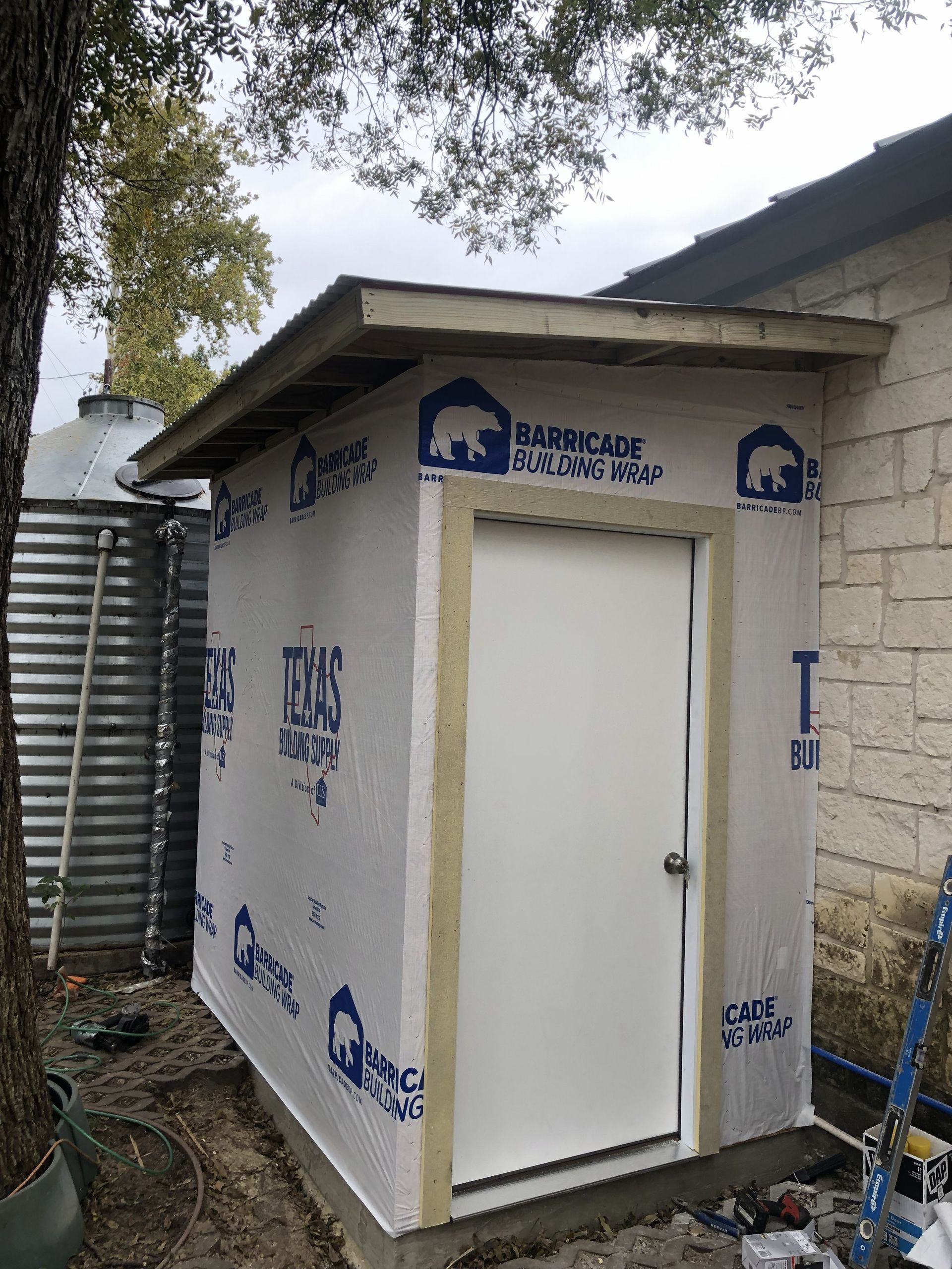 Small shed with a white door, partially covered in blue building wrap. Next to a metal water tank.