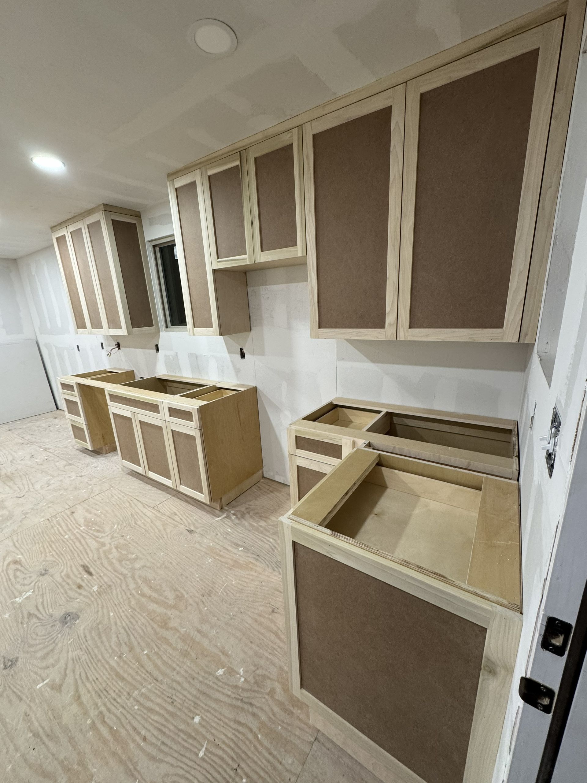 Kitchen cabinets under construction, unfinished wood, neutral tones, mounted on walls, with some lower units.