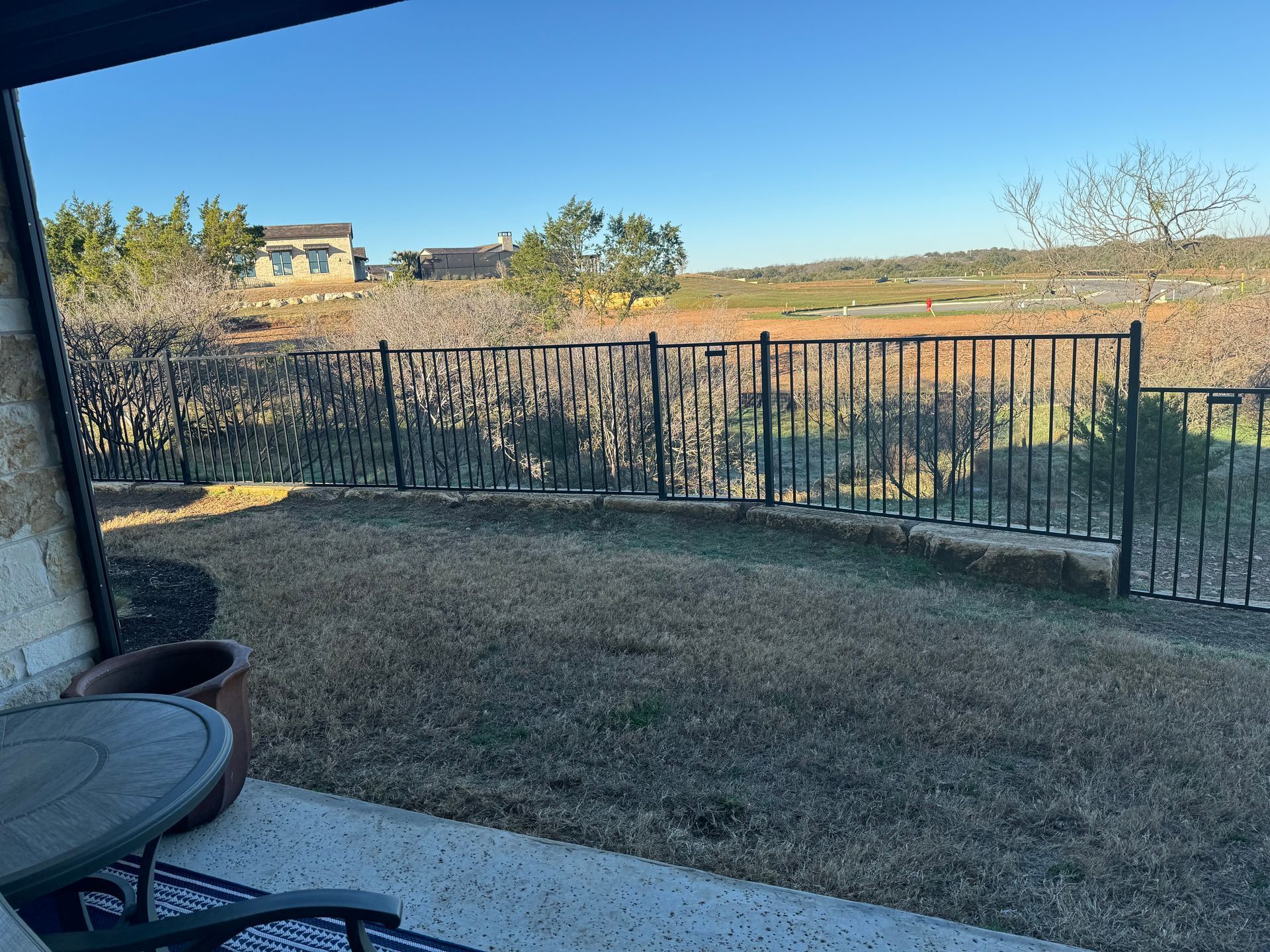 Patio with a metal fence overlooking a dry, grassy field under a blue sky.