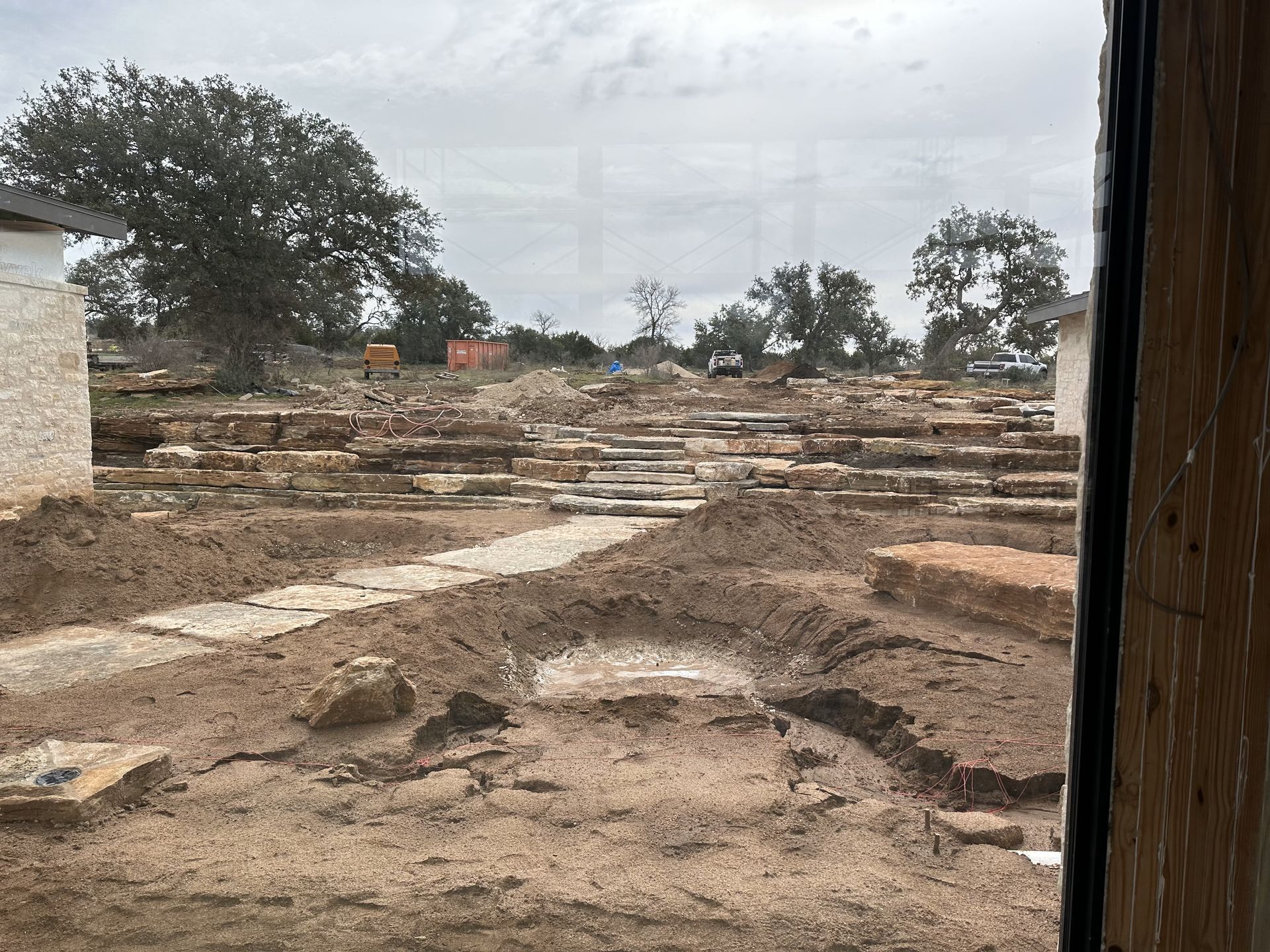 Construction site with exposed dirt and stone foundations. Cloudy day, trees in background.