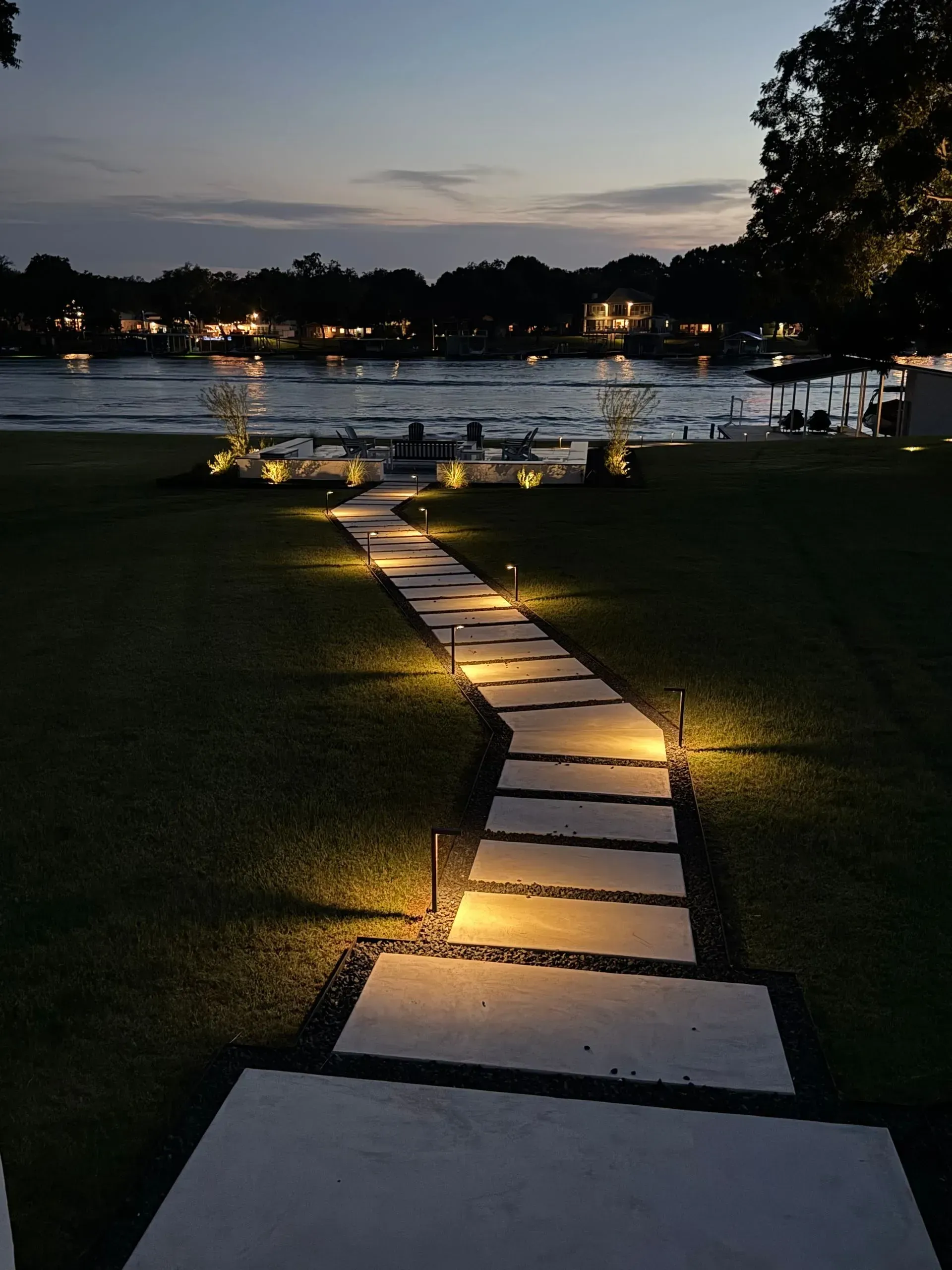 A stone pathway illuminated by lights leads to a waterfront at dusk.