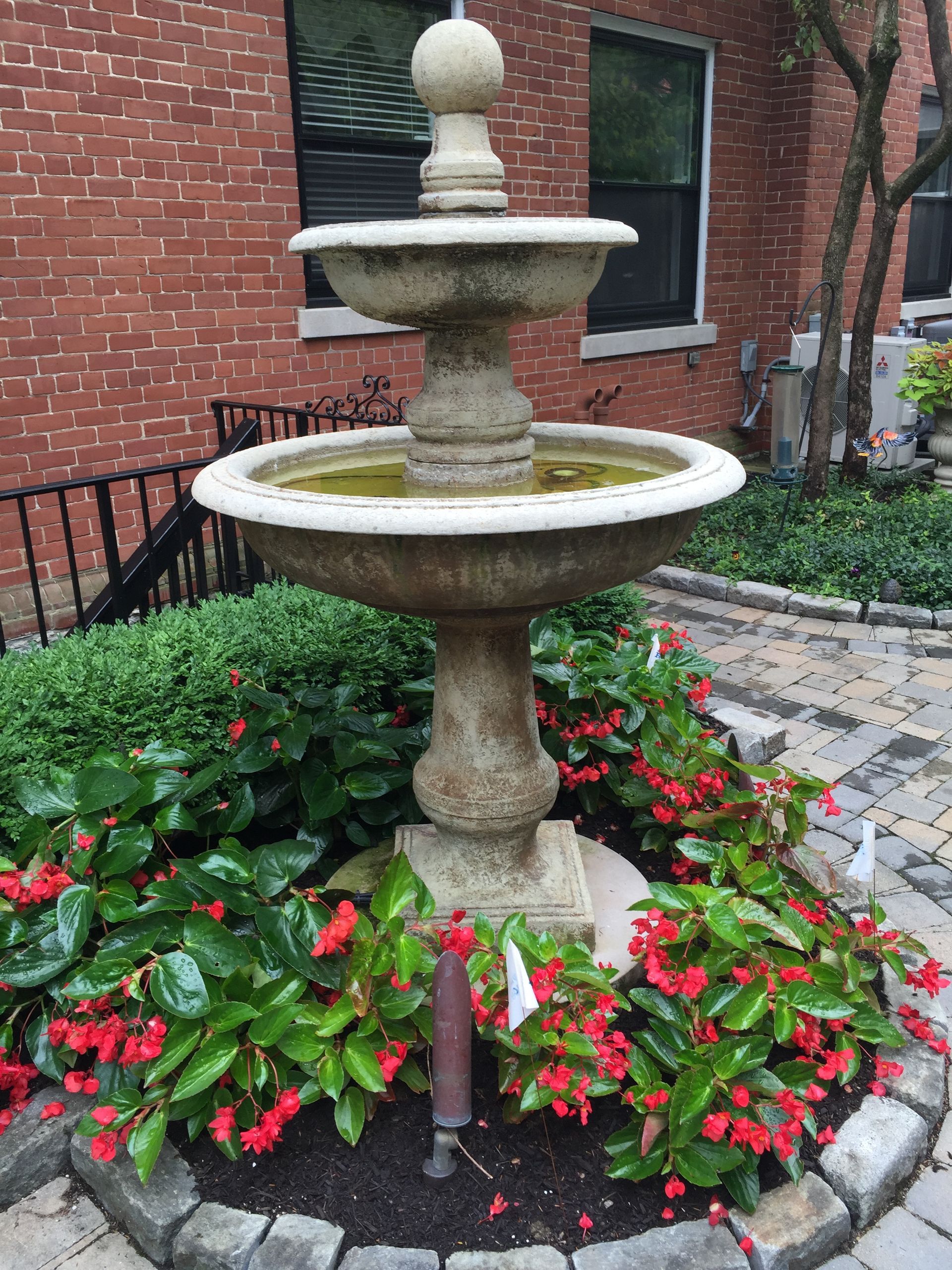 A fountain in a garden with red flowers in front of a brick building