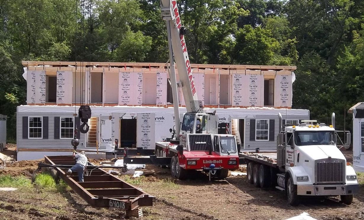 A crane lifts a modular home section into place at a construction site near a forest.