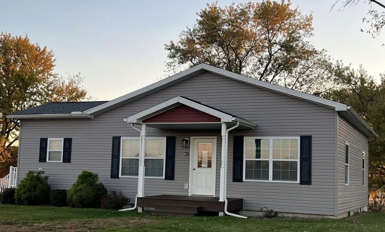 Single-story gray house with a small covered entryway, white door, dark shutters, and autumn trees in the background.