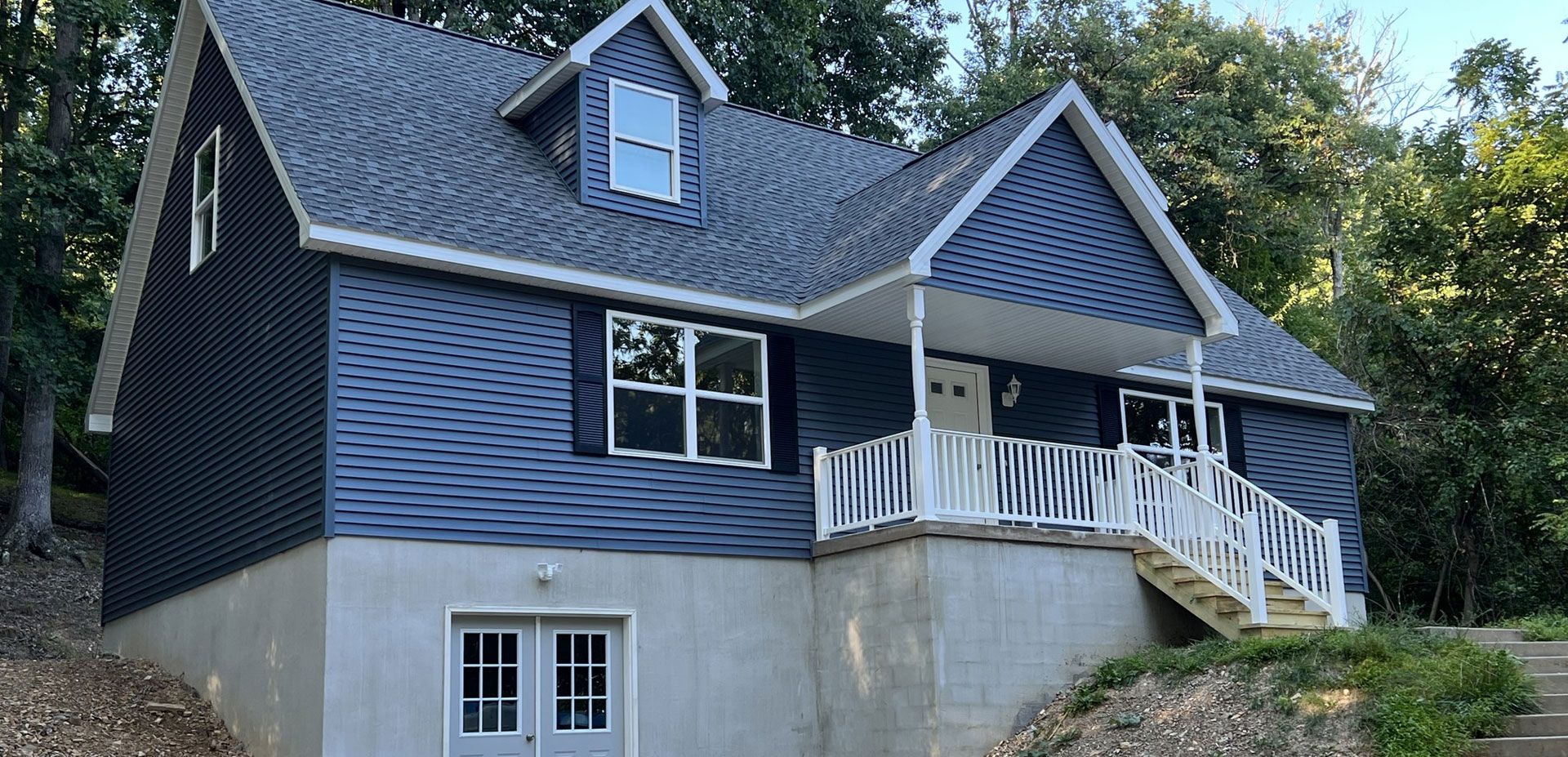 A two-story blue house with a white front porch and a basement-level set of French doors, nestled in a wooded area.