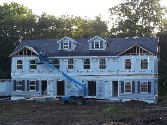 A two-story building under construction with a blue boom lift parked in front against a wooded background.