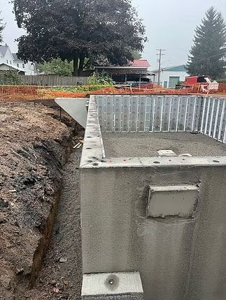 A partially constructed concrete foundation wall in an excavated dirt site, with a drainage trench and gravel base.