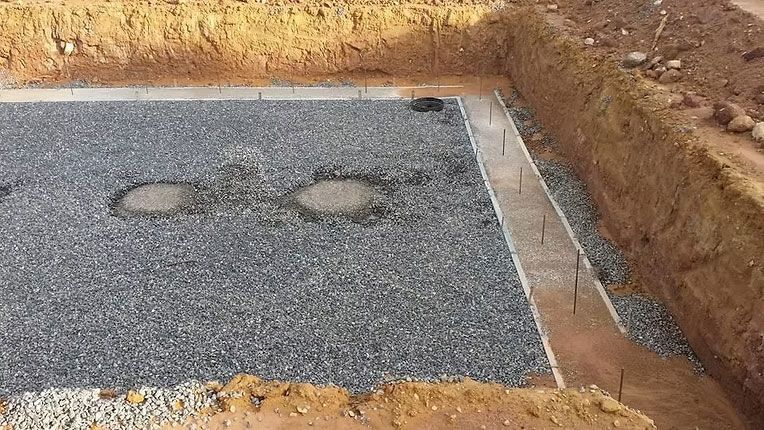 A construction site excavation featuring a gravel-filled foundation area with perimeter concrete footings and metal stakes.