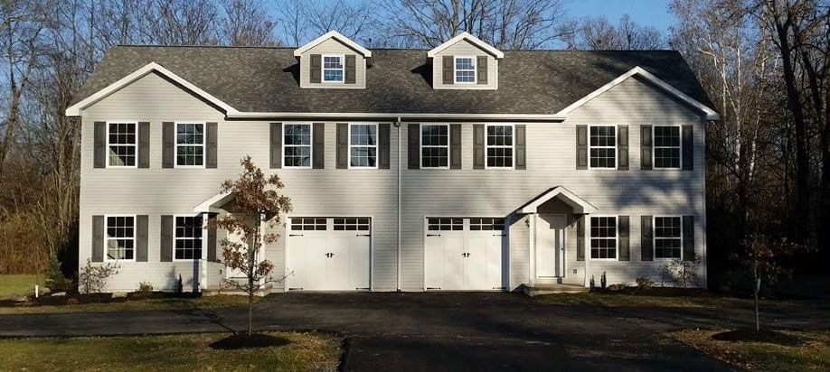 A two-story duplex with light gray siding, dark shutters, two garage doors, and two small dormer windows.