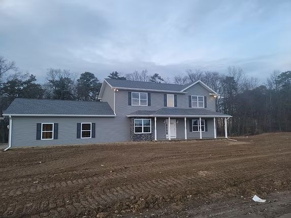 A newly constructed gray, two-story house with a covered front porch, set on a bare dirt lot under a cloudy sky.