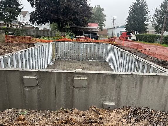 An excavated pool site with grey concrete foundation walls and metal supports on a dirt lot in a residential area.