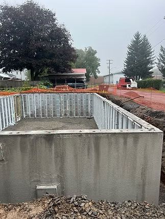 A concrete foundation for a new structure set in an outdoor yard with trees and a red construction path in the background.