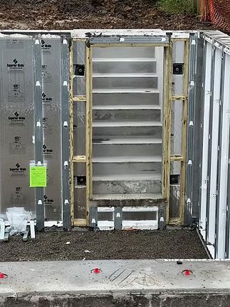 A construction site showing concrete foundation walls and a wooden framed doorway leading to interior concrete stairs.