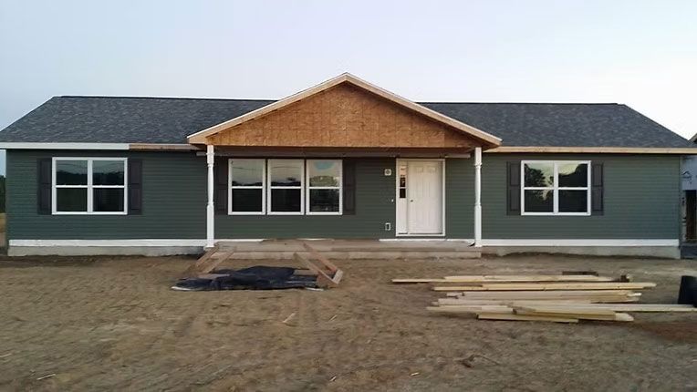 A single-story house under construction with dark green siding, white trim, a gabled porch entrance, and a dirt yard.