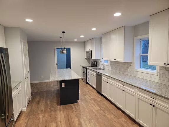 A modern kitchen features white cabinets, gray granite countertops, a dark island, and light wood-look flooring.