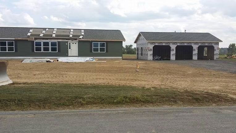 A newly constructed olive-green ranch-style home sits next to an unfinished detached garage on a dirt lot.