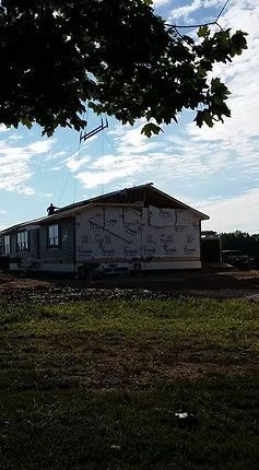 A house under construction with exposed sheathing, framed against a cloudy blue sky under the branches of a large tree.