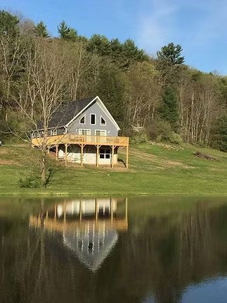 A grey two-story house with a wooden deck overlooks a calm lake, reflecting perfectly on the water against a hillside.